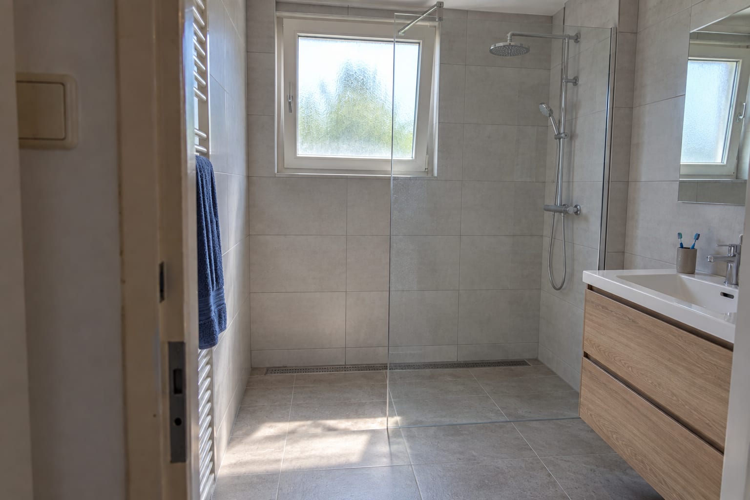 Na de verbouwing: een moderne badkamer met een inloopdouche achter glas en grote grijze tegels