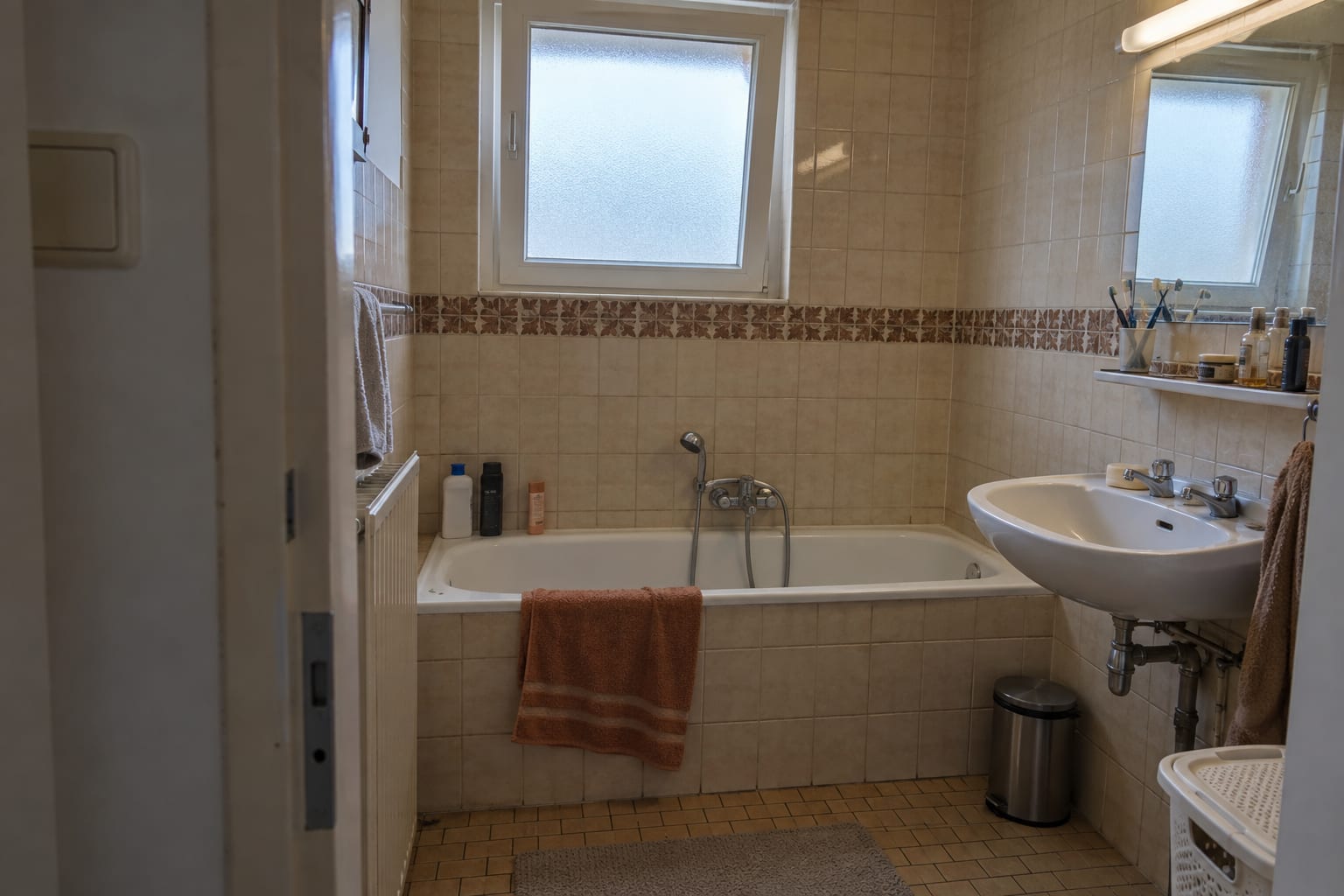 Voor: gedateerde badkamer met beige tegels, een ligbad en losse kranen